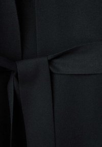 Gros plan d'une ceinture en tissu noire nouée d'un simple nœud autour d'un vêtement, montrant la texture tricotée et la finition lisse.