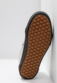 Sneaker-Sohle mit einer strukturierten Gummioberfläche, die ein Diamantmuster aufweist. Das Obermaterial ist aus leicht beigem Material mit dunklen Akzenten.