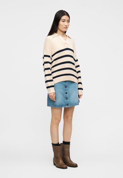 Γυναίκα που φοράει μπεζ πουλόβερ με μαύριες ρίγες, τζιν φούστα με κουμπιά στο μπροστινό μέρος, μαύρες κάλτσες και καφέ μπότες μέχρι τη μέση της γάμπας, στέκεται σε λευκό φόντο.