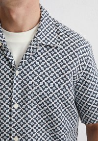 Camicia a maniche corte con motivo geometrico in navy e azzurro chiaro su sfondo bianco. Include una tasca sul petto e chiusura con bottoni.