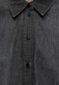 Gros plan d'une chemise en denim gris foncé avec un col pointu et quatre boutons noirs sur le devant.