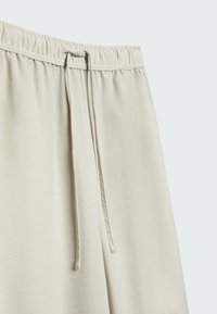 Gros plan sur un pantalon beige avec une ceinture élastique et un cordon de serrage au centre sur un fond clair.