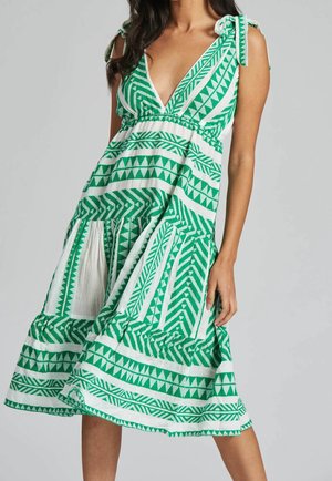 Kobieta w białej, bezrękawnikowej sukience do kolan z zielonymi geometrycznymi wzorami i wiązanymi ramiączkami, stojąca na jednolitym tle.