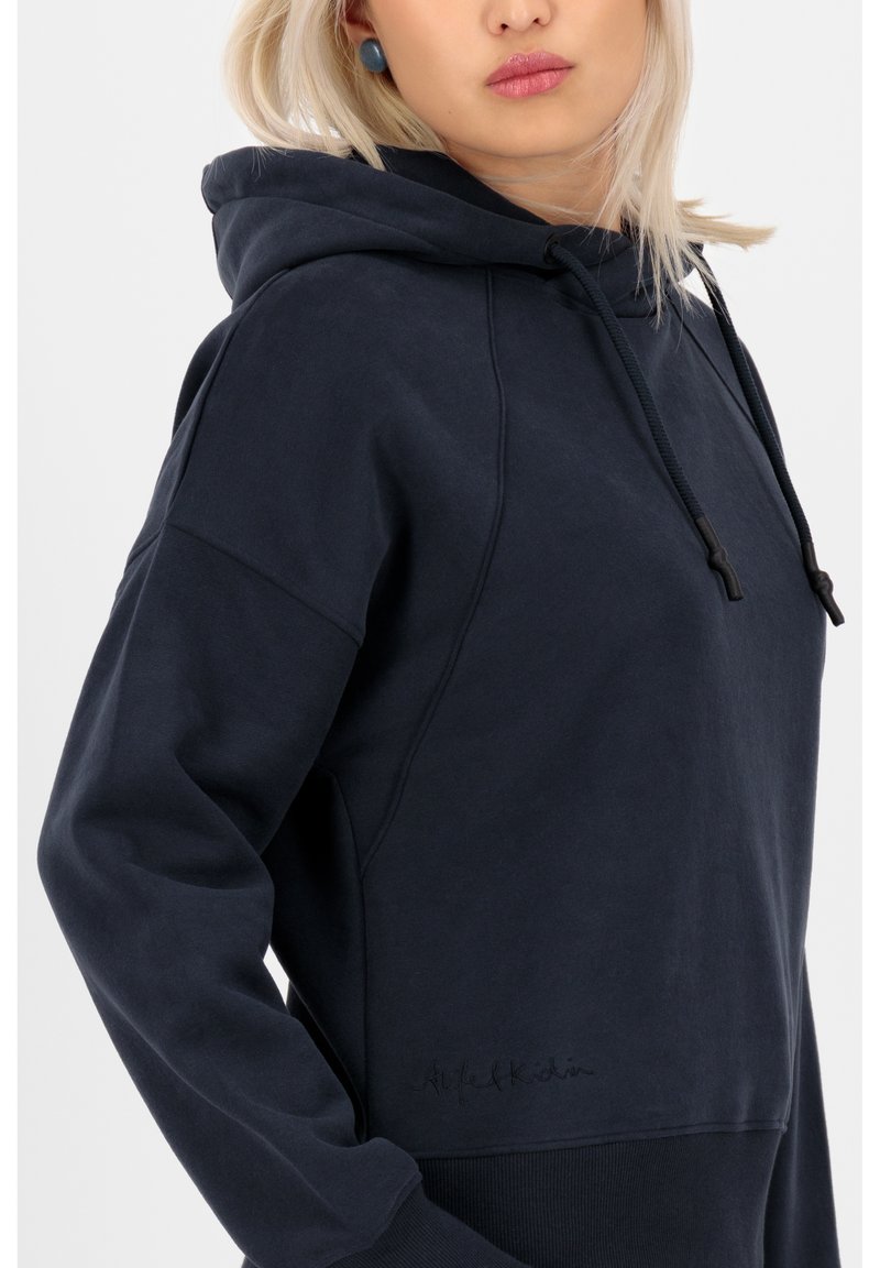 Donna con capelli biondi che indossa una felpa con cappuccio blu scuro con cordini, mostrata dalla spalla a metà torso su uno sfondo bianco.