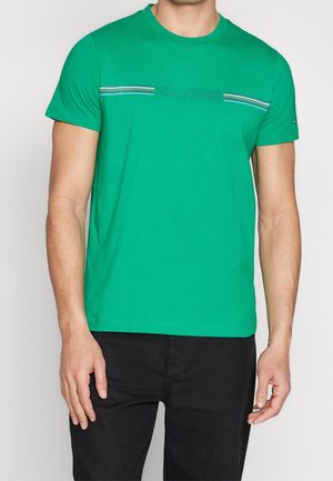 Homme portant un t-shirt vert vif à manches courtes avec des rayures horizontales et le texte « HILFIGER », assorti à un pantalon noir.