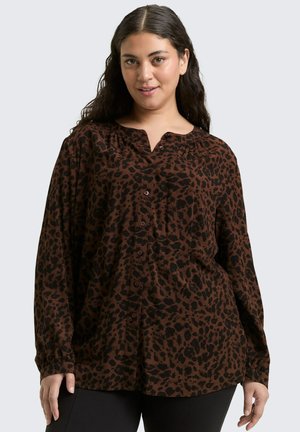 Femme avec de longs cheveux noirs portant une blouse à motifs léopard marron et noir, ample, avec des boutons, et un pantalon noir, souriant légèrement.
