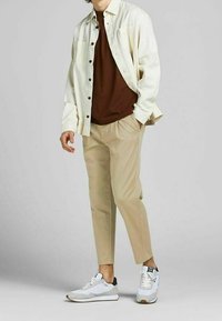 Vit skjorta med knappar över en brun t-shirt, beige byxor och grå sneakers. Funktioner inkluderar två bröstfickor och kontrasterande skofärger.