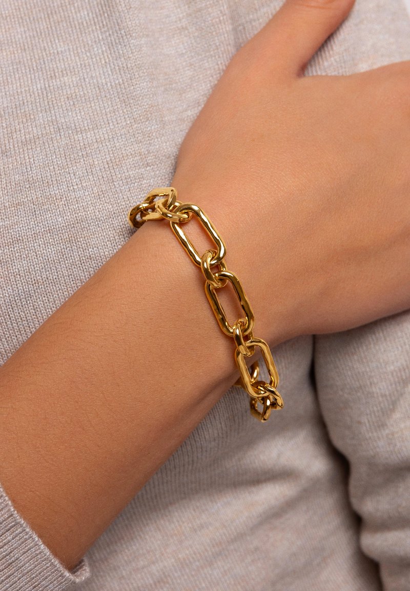Guldkedja armband med stora, texturerade länkar i en avlång, rektangulär form. Bärs på ett handled mot en ljus, texturerad bakgrund.