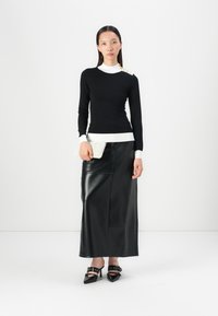 Zwarte langarmtop met een witte kraag en gouden knoopdetails, gecombineerd met een zwarte leren maxi rok en zwarte puntige hakken. Houdt een witte clutch vast.