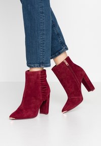 Stivaletti in camoscio bordeaux con punta affusolata, tacco a blocco e dettagli arricciati, dotati di una zip laterale in una finitura metallica a contrasto.