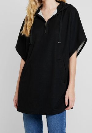 Vrouw draagt een zwart kortewerps capuchontrui met rits aan de voorkant en kangoeroezak, gecombineerd met blauwe spijkerbroek.