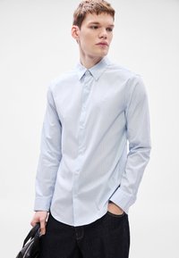 Camisa de rayas azul claro con botones, cuello estándar, mangas largas y dobladillo redondeado, combinada con pantalones vaqueros oscuros y un bolso negro.