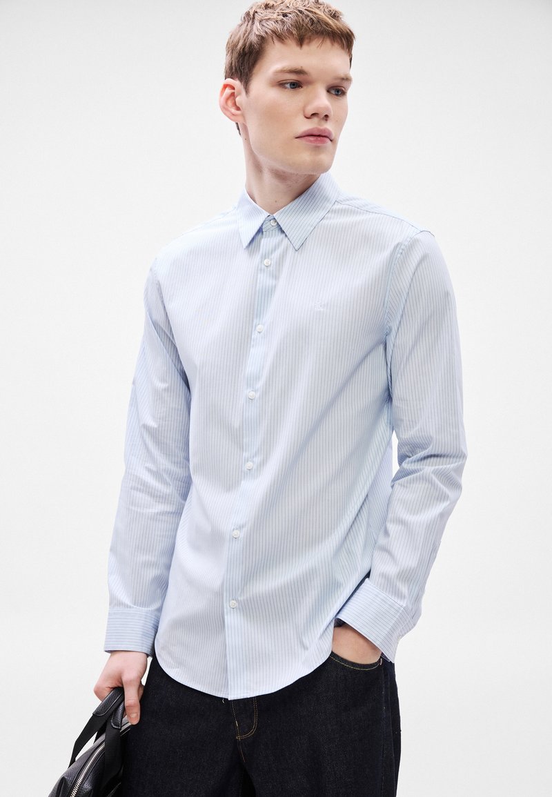 Hellblau gestreiftes Hemd mit Knöpfen, Standardkragen, langen Ärmeln und abgerundetem Saum, kombiniert mit dunklen Jeanshosen und einer schwarzen Tasche.