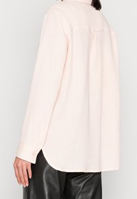 Mujer vestida con una camiseta de manga larga texturizada en rosa claro y pantalones de cuero negros, vista desde la espalda y el costado.