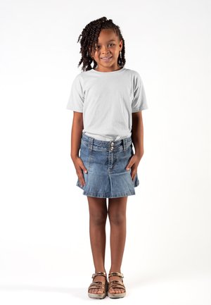 Giovane ragazza con i capelli intrecciati che indossa una maglietta bianca, una gonna di jeans e sandali dorati, sorridente mentre è in piedi davanti a uno sfondo bianco.