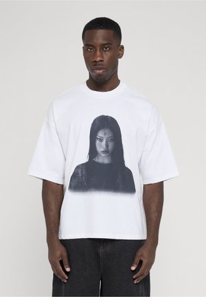Mand står mod en ensfarvet baggrund iført en hvid T-shirt med et sort-hvidt portræt af en kvinde trykt på forsiden.