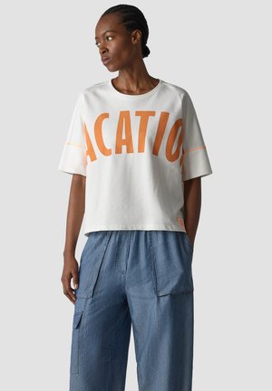 Woman with braided hair wearing a white oversized T-shirt with large orange text and loose blue denim cargo pants against gray background.