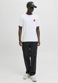 Hvid bomulds t-shirt med et lille rødt firkantet logo, parret med mørke denim jeans og lysegrå sneakers. En sort kasket fuldender looket.
