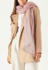 Écharpe légère rose pâle avec des bords à franges, drapée de manière décontractée sur un trench beige et un haut blanc. Texture lisse, tissu doux.
