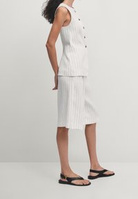 Wit gestreept pakken set met een mouwloze top met knopen en zakken, gecombineerd met knielange shorts en zwarte platte sandalen.
