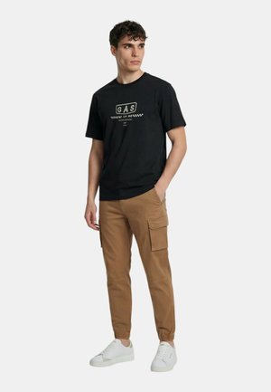 Giovane uomo con indosso una T-shirt nera con grafica, pantaloni cargo marroni e scarpe da ginnastica bianche, in piedi con una mano in tasca, guarda di lato.