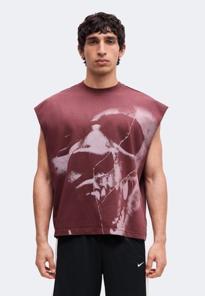 Jeune homme portant un débardeur bordeaux avec un grand motif de crâne délavé et un short Nike noir, debout devant un fond uni.