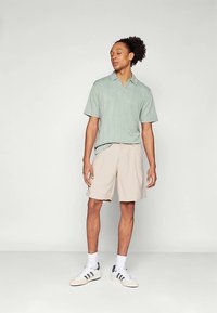 Ανοιχτό πράσινο κοντομάνικο πόλο με γιακά, φορεμένο με μπεζ σορτς και λευκά αθλητικά με μαύρες ρίγες. Λείο ύφασμα, άνετη εφαρμογή.