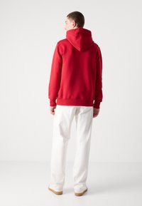 Sudadera roja con capucha, con un bolsillo tipo canguro, puños y dobladillo acanalados. Se lleva con pantalones blancos y zapatos beige. Fondo liso.