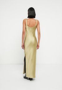 Μακριά, εφαρμοστή φόρεμα σε φωτεινό ελαιόχρωμο σατέν με στρογγυλή πλάτη, πλαϊνή σχισμή και λεπτές τιράντες· συνοδευόμενο από μαύρα μποτάκια με τακούνι.