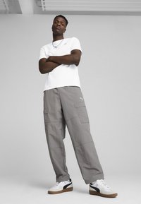 Hvid t-shirt med korte ærmer, grå cargo-bukser og hvide sneakers med sorte detaljer. Model står med armene over kors foran en lys baggrund.