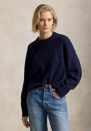 Jeune femme aux cheveux blonds courts portant un pull bleu foncé et un jean bleu, debout avec une main dans la poche devant un fond uni.