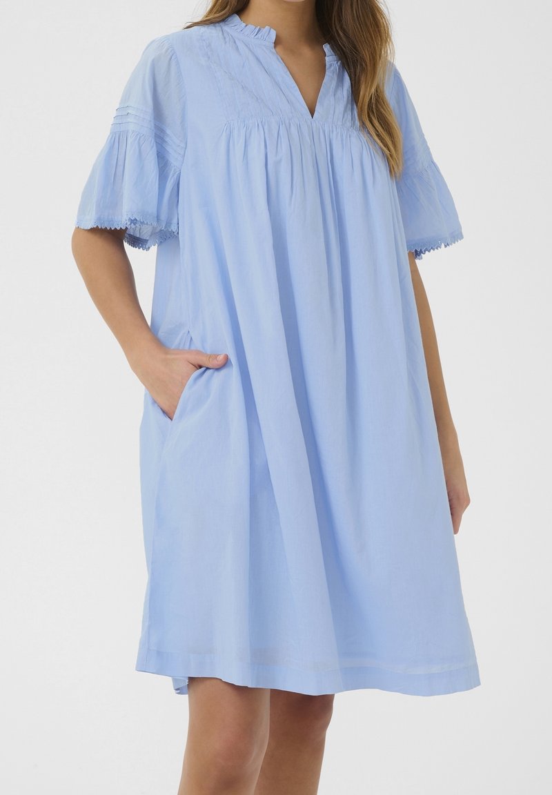 Ljusblå bomullsklänning med V-ringning, korta puffärmar med spetsdetaljer och sidofickor. Designen är löst sittande.