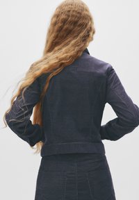 Jachetă din denim de culoare albastră închisă, cu un design ajustat, cusături vizibile pe piept și pe laterale, și mâneci lungi. Țesătură texturată cu detalii minime.