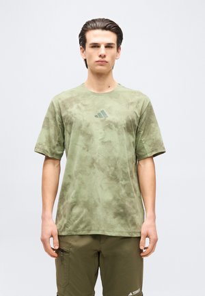 Noor mees, kes kannab oliivrohelist tindivärvitud t-särki logoga ja sobivaid tehnikapükse, seisab ühevärvilise hele tausta ees.