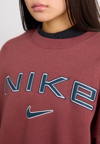 Bordový sweatshirt se velkým bílým a modrým vyšívaným logem "NIKE" a malým modrým swoosh. Hladký černý límec. Měkký bavlněný materiál.