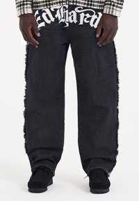 Jeans in denim nero con dettagli sfilacciati lungo i lati e grande testo bianco "Ed Hardy" sulla cintura anteriore. Design morbido e rilassato.