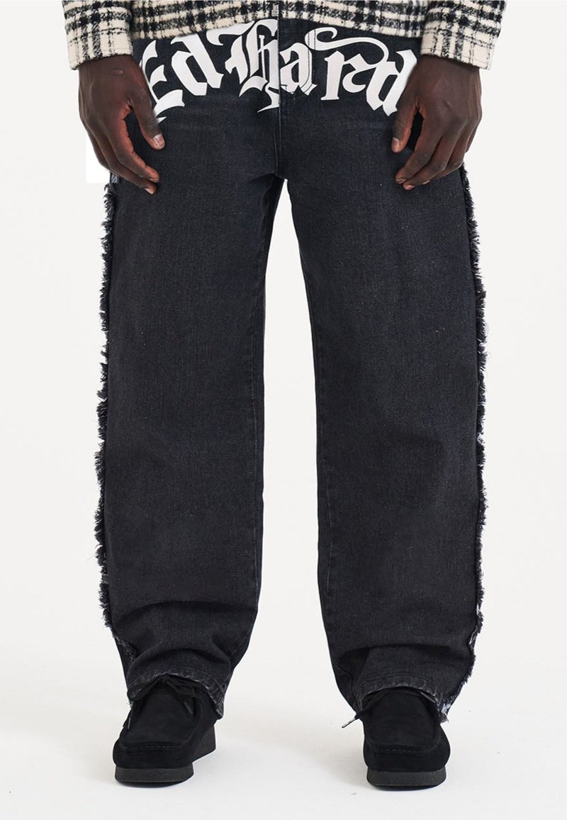Jeans in denim nero con dettagli sfilacciati lungo i lati e grande testo bianco "Ed Hardy" sulla cintura anteriore. Design morbido e rilassato.