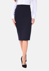 Gonna a matita blu navy, lunghezza al ginocchio, con vita elastica e design aderente. Indossata con una camicia bianca a maniche lunghe e tacchi neri su uno sfondo neutro.