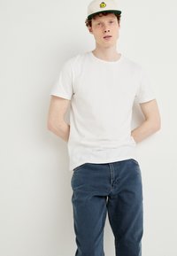 Camiseta de algodão branca com decote redondo, combinada com jeans de denim azul escuro. Um boné bege com aba verde e detalhes do logótipo amarelo está sendo usado.