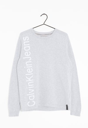 Jasnoszara bluza z długim rękawem z napisem "Calvin Klein Jeans" dużymi białymi pionowymi literami, wisząca na czarnym wieszaku na białym tle.