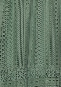 Tissu en dentelle verte présentant des motifs complexes, y compris des formes circulaires et en losange, avec une finition texturée et un design superposé.