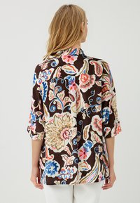 Kobieta z blond włosami, nosząca luźną, kolorową bluzkę w kwiatowy wzór z rękawami 3/4, widziana z tyłu na jednolitym tle.