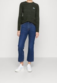 Ciemnozielony, długi sweter z dzianiny z długim rękawem, okrągłym dekoltem i małym logo. Połączony z wysokim stanem, niebieskimi, krótkimi dżinsami i białymi trampkami.
