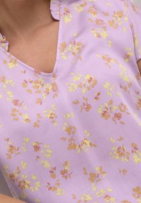 Fialová blůza s výstřihem do V a volánkovým límcem, zdobená malými, rozptýlenými květinovými vzory v žlutých a jemně růžových tónech.