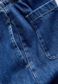 Džínové nohavice tmavomodrej farby s predným vreckom, elastickým pásom a viditeľnými detailmi švov. Hladká textúra.