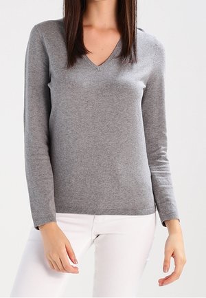 Kvinde iført en tætsiddende grå V-hals trøje med lange ærmer og hvide bukser, stående foran en ensartet lys baggrund.