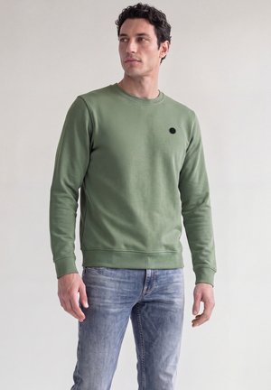 Homme portant un sweat-shirt vert à col rond et un jean bleu délavé, debout devant un fond clair uni, regardant sur le côté.