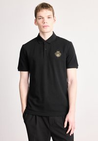 Melna kokvilnas polokrāsa ar īsām piedurknēm. Zeltītā izšūtā logo kreisajā krūtīs. Parasts apkakles dizains.