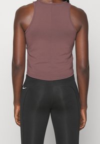 Personne portant un haut court sans manches mauve et des leggings Nike noirs, vue de dos sur un fond clair uni.