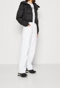 Doudoune noire avec capuche, fermeture à zip et poignets élastiques, associée à un jean blanc à jambes larges et des chaussures de sport noires.